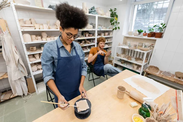 Joyful and interracial man and woman in aprons handcrafting during pottery lesson — Stock Photo