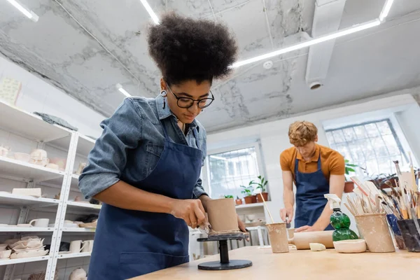 Curly african american woman in eyeglasses molding clay cup near blurred and redhead man — Stock Photo