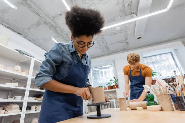 Frisé afro-américain femme dans lunettes moulage tasse d'argile près floue et rousse homme — Photo de stock