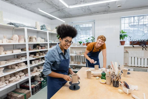 Gai afro-américaine femme en lunettes moulage tasse d'argile près de rousse homme — Photo de stock