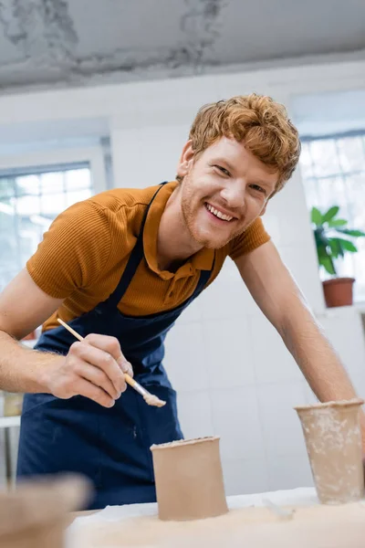 Cheerful and redhead man in apron holding shaper near clay cup while looking at camera — Stock Photo