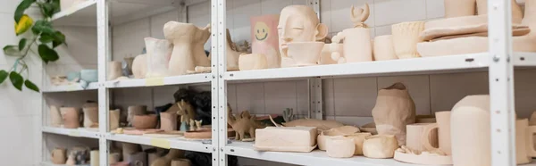 Different clay sculptures near cups and bowls on shelves of pottery shop, banner — Stock Photo