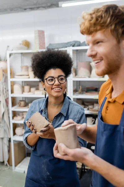 Feliz africana americana mujer mirando pelirroja novio moldeado arcilla taza en cerámica estudio - foto de stock