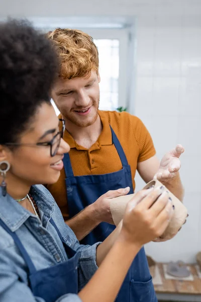 Happy redhead man pointing with hand at clay cup during date with african american girlfriend in pottery studio — Stock Photo