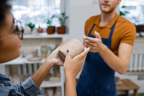 Homme barbu pointant du doigt la tasse d'argile dans les mains de la femme afro-américaine en atelier de poterie — Photo de stock