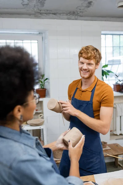 Happy redhead man molding clay cup during date with african american girlfriend in pottery studio — Stock Photo
