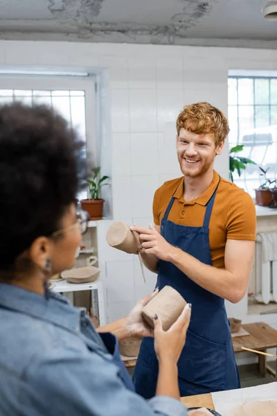 Heureux rousse homme moulage tasse d'argile pendant la date avec petite amie afro-américaine en studio de poterie — Photo de stock