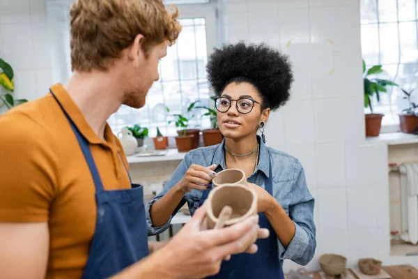 Lockige afrikanisch-amerikanische Frau mit Brille schaut den rothaarigen Freund an, der im Töpferatelier Tonbecher gießt — Stockfoto