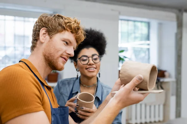 Barbudo pelirrojo hombre comprobar arcilla taza cerca feliz africano americano novia en gafas - foto de stock