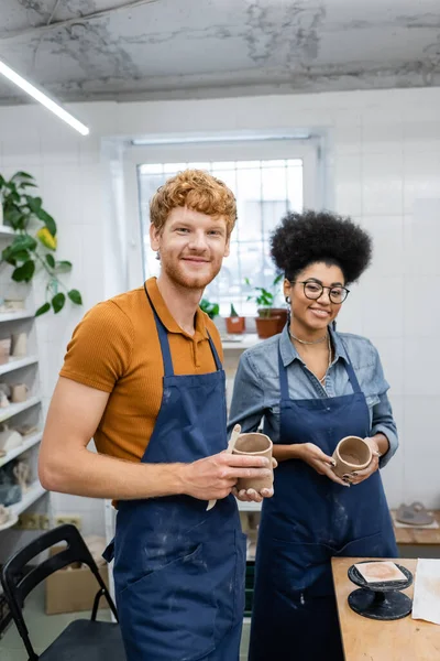 Alegre casal multiétnico em aventais segurando copos de barro no estúdio de cerâmica — Fotografia de Stock