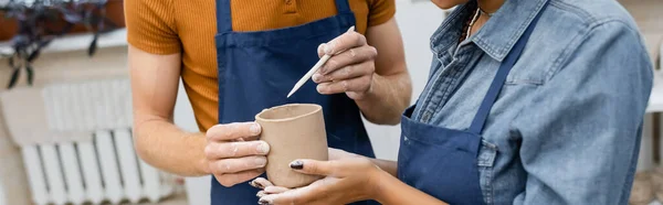 Vue recadrée de femme afro-américaine près de l'homme en tablier modèle tasse d'argile avec shaper, bannière — Photo de stock