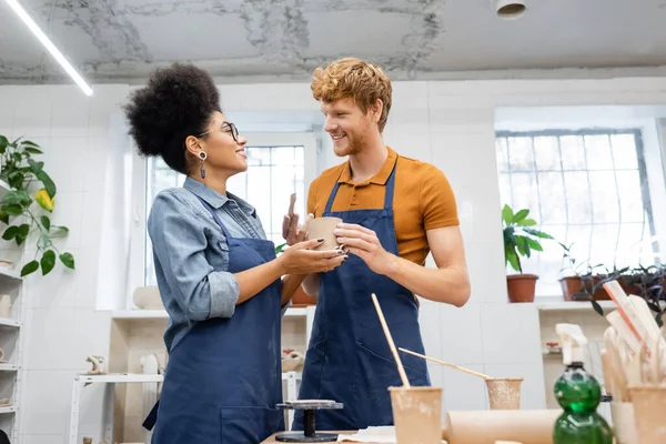 Feliz pareja multiétnica en delantales mirándose mientras sostiene la taza de arcilla en el estudio de cerámica - foto de stock