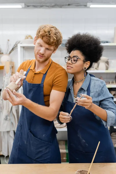 Artigiano in possesso di strumento e argilla vicino donna afro-americana in laboratorio di ceramica — Foto stock