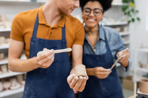 Profesor borroso sosteniendo escultura de arcilla y formador cerca de mujer afroamericana en taller — Stock Photo