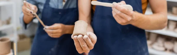 Vue recadrée de l'artisan tenant le shaper et le produit d'argile près de femme afro-américaine floue dans l'atelier, bannière — Photo de stock