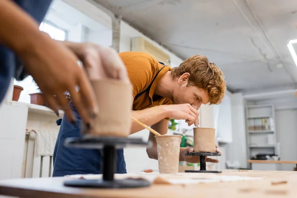 Pelirroja hombre formando escultura de arcilla cerca borrosa novia afroamericana en estudio de cerámica - foto de stock