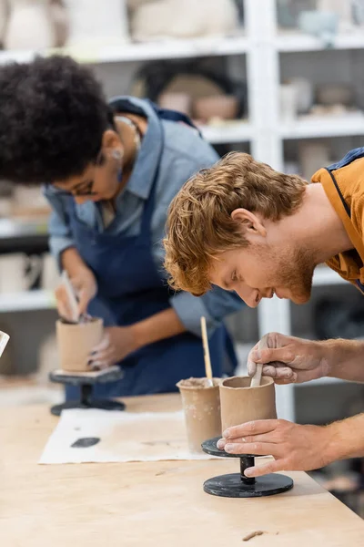 Smiling man forming clay sculpture near blurred african american girlfriend in pottery studio — Stock Photo