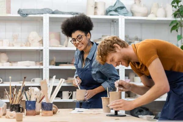 Femme afro-américaine souriante dans un tablier tenant de l'argile près de son petit ami dans un studio de poterie — Photo de stock