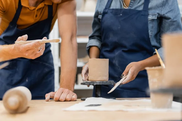 Ausgeschnittene Ansicht eines Handwerkers mit Werkzeug in der Nähe einer afrikanisch-amerikanischen Frau und Skulptur in einer Töpferei-Werkstatt — Stockfoto