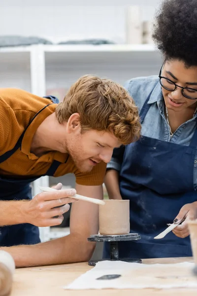 Smiling redhead teacher holding tool near clay cup and african american woman in pottery workshop — Stock Photo