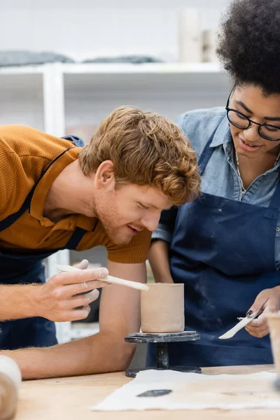 Lächelnde rothaarige Lehrerin hält Werkzeug in der Nähe von Tonbecher und afrikanisch-amerikanische Frau in Töpferwerkstatt — Stockfoto