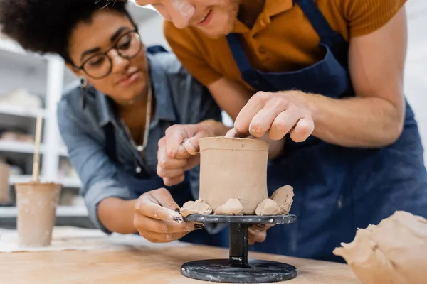 Hombre sonriente formando escultura de arcilla cerca de borrosa profesora afroamericana en taller - foto de stock