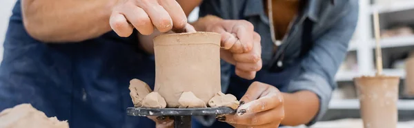 Vista recortada del hombre haciendo escultura de arcilla cerca de artesana afroamericana en taller de cerámica, pancarta - foto de stock