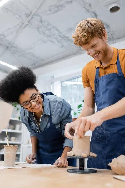 Smiling man making ceramic cup near african american craftswoman in pottery studio — Stock Photo