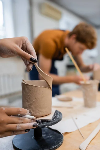 African american woman holding tool and clay near blurred boyfriend during date in pottery studio — Stock Photo