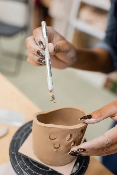 Ausgeschnittene Ansicht einer afrikanisch-amerikanischen Kunsthandwerkerin, die Werkzeug in der Nähe von Tonprodukten in der Werkstatt hält — Stockfoto