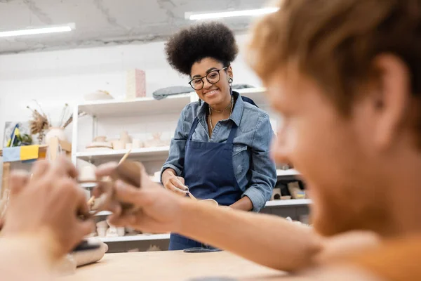 Smiling african american woman in apron looking at blurred boyfriend during date in pottery workshop — Stock Photo