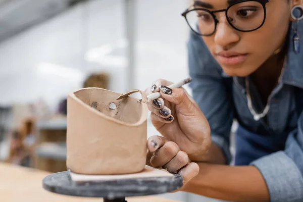 Artesano afroamericano borroso en anteojos cortando escultura de arcilla en taller - foto de stock