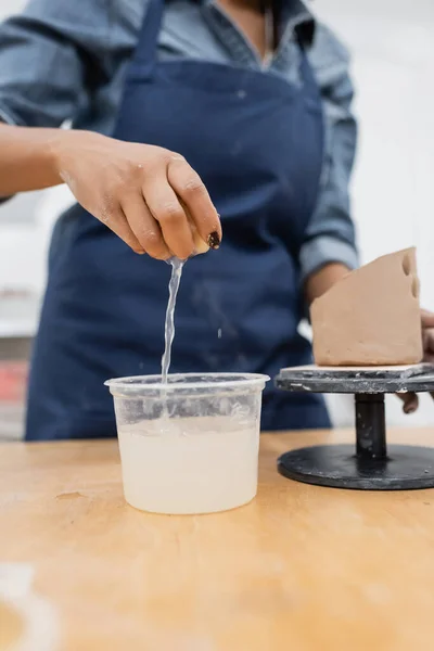 Ausgeschnittene Ansicht einer afrikanisch-amerikanischen Kunsthandwerkerin, die Schwamm mit Wasser in der Nähe von Ton im Töpferatelier hält — Stockfoto