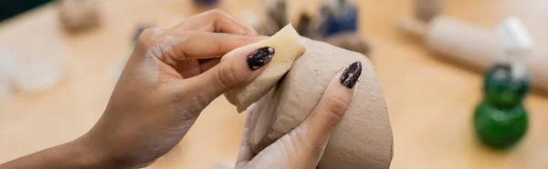 Vista recortada de artesano afroamericano sosteniendo esponja húmeda y arcilla en taller de cerámica, pancarta - foto de stock