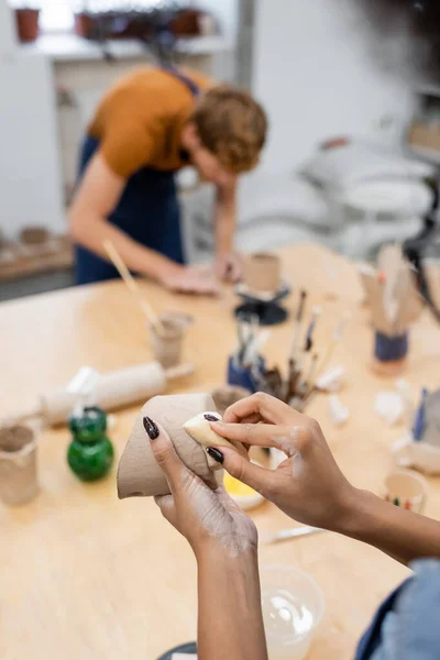 Donna afroamericana in possesso di spugna e argilla vicino fidanzato offuscata in laboratorio di ceramica — Foto stock