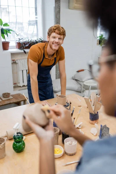 Sorrindo homem olhando para a namorada afro-americana borrada perto de barro no estúdio de cerâmica — Fotografia de Stock