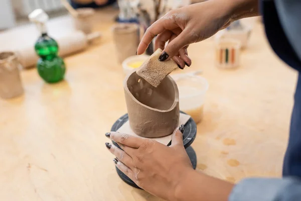Vista cortada de Africano americano mestre segurando esponja perto de produto de argila em oficina de cerâmica — Fotografia de Stock