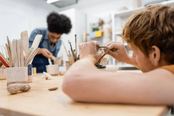 Rotschopf-Mann formt Tonbecher in der Nähe von Werkzeugen und verschwommener afrikanisch-amerikanischer Freundin im Töpferstudio — Stockfoto