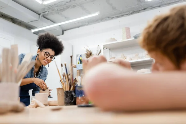 Sourire femme afro-américaine tenant de l'argile près des outils flous et petit ami dans un atelier d'argile — Photo de stock