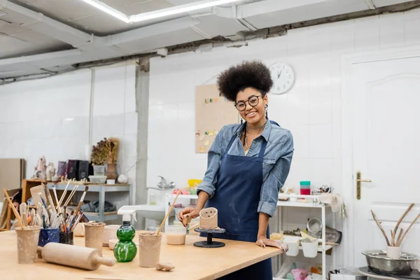 Happy african american artisan in apron looking at camera near clay and tools in pottery studio — Stock Photo