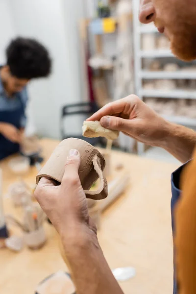 Vista ritagliata dell'uomo che tiene la tazza di argilla e la spugna durante la data con la fidanzata africana americana offuscata nel laboratorio di ceramica — Foto stock