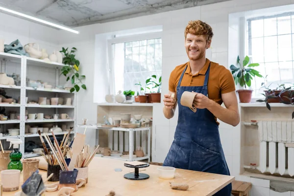 Sourire rousse artisan dans le tablier regardant la caméra tout en faisant tasse en céramique dans l'atelier — Photo de stock