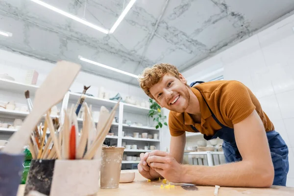 Artisanat rousse souriant regardant la caméra près des outils de poterie flous dans l'atelier — Photo de stock