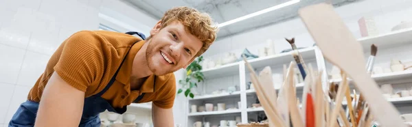 Joyeux artisan regardant la caméra près des outils de poterie flous dans l'atelier, bannière — Photo de stock