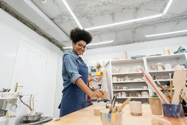 Young african american craftswoman smiling at camera near clay and tools in pottery workshop — Stock Photo