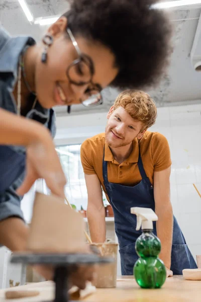 Smiling man in apron looking at blurred african american girlfriend making clay sculpture in pottery studio — Stock Photo