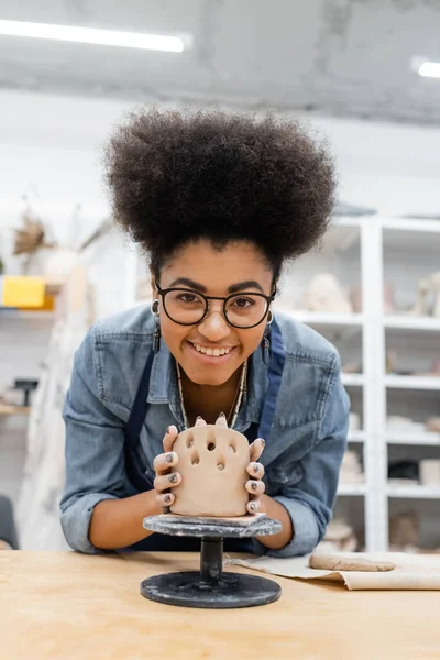 Cheerful african american craftswoman in eyeglasses making ceramic product in pottery workshop — Stock Photo