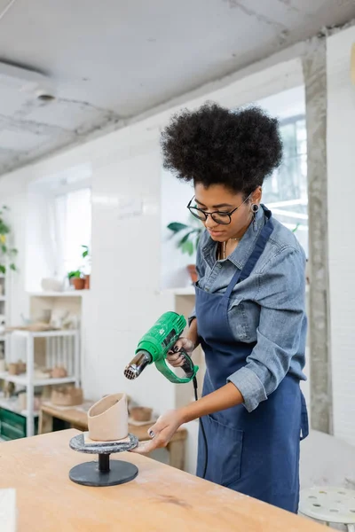 Artesana afroamericana sosteniendo pistola de calor cerca de escultura de arcilla en taller de cerámica - foto de stock