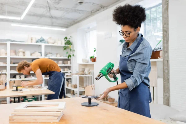 Mujer afroamericana sosteniendo pistola de calor cerca de producto de arcilla y novio borroso en taller de cerámica - foto de stock