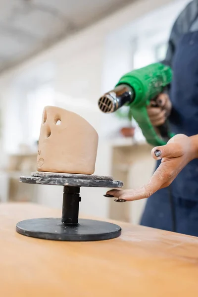 Vista recortada de artesano afroamericano sosteniendo pistola de calor borrosa cerca de escultura de arcilla en taller de cerámica - foto de stock